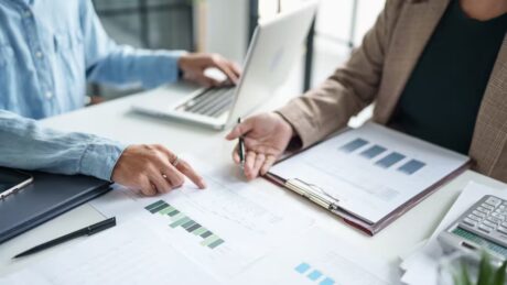 two people sitting at a desk looking at projected sales numbers on sheets of paper